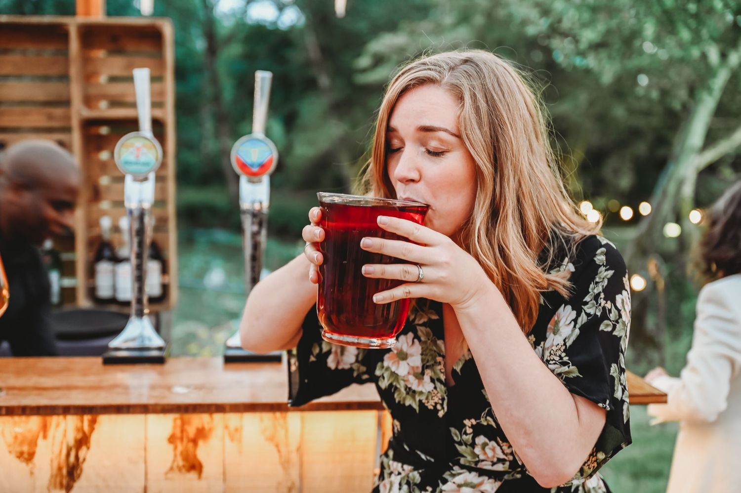 A woman in a floral dress sips a red beverage while sitting at an outdoor bar with string lights in the background.