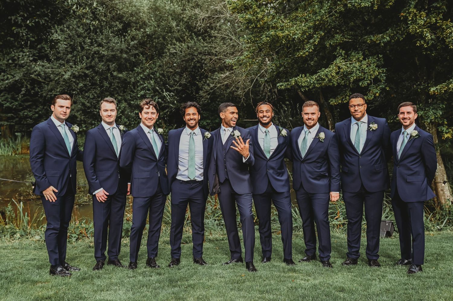 A groomsmen party in navy suits stand together for a photo on a green lawn.