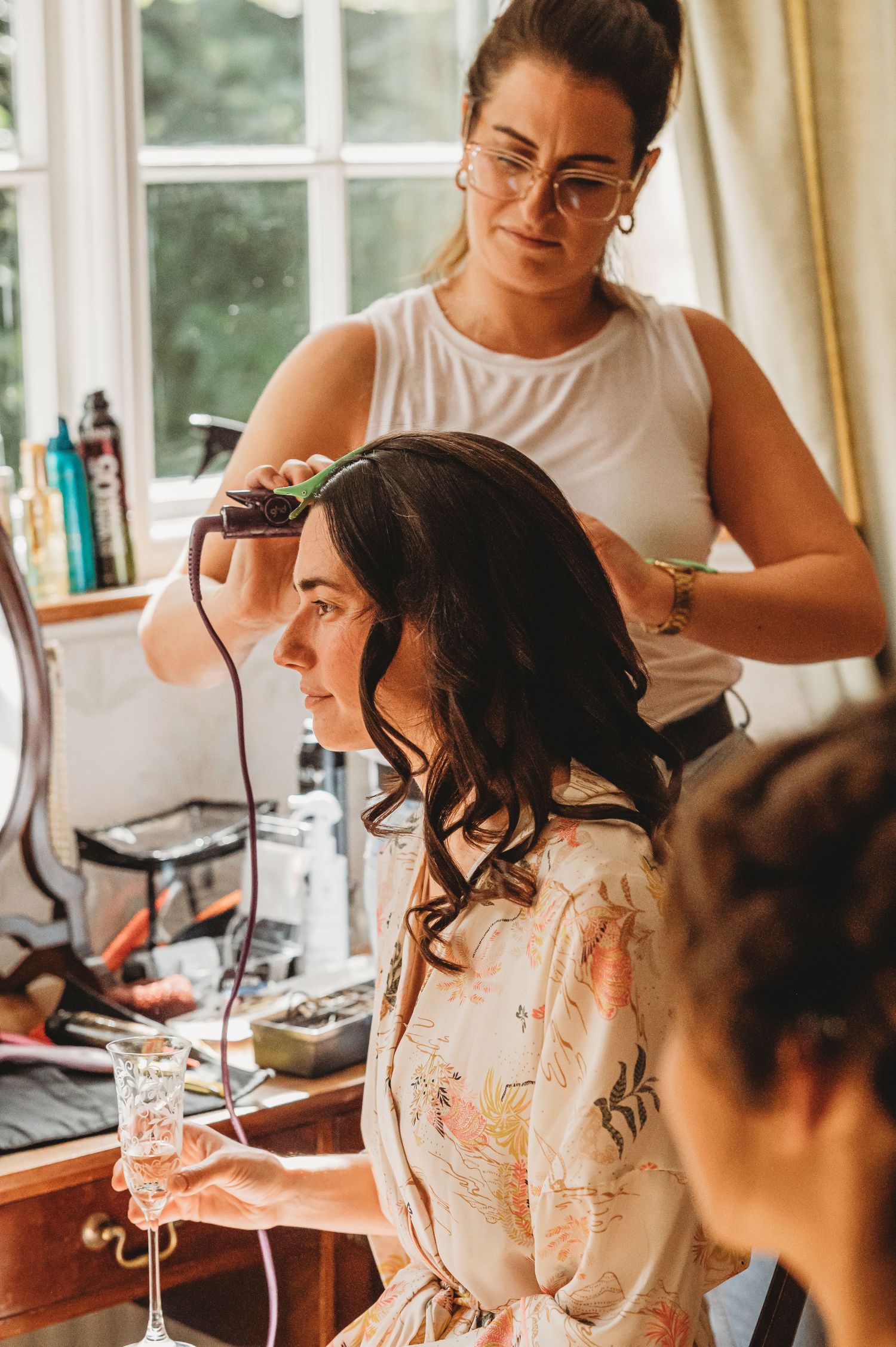 Hair stylist using curling iron to style client's dark wavy hair during bridal preparation in sunlit room.