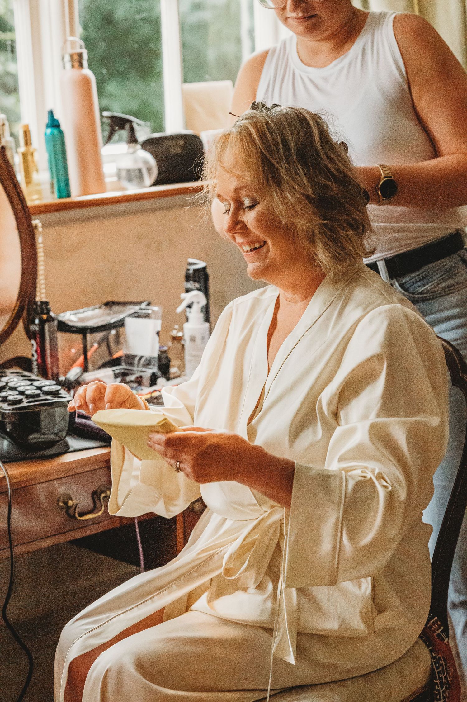 Someone gets their hair styled while wearing a white robe and sitting at a makeup station with beauty products.