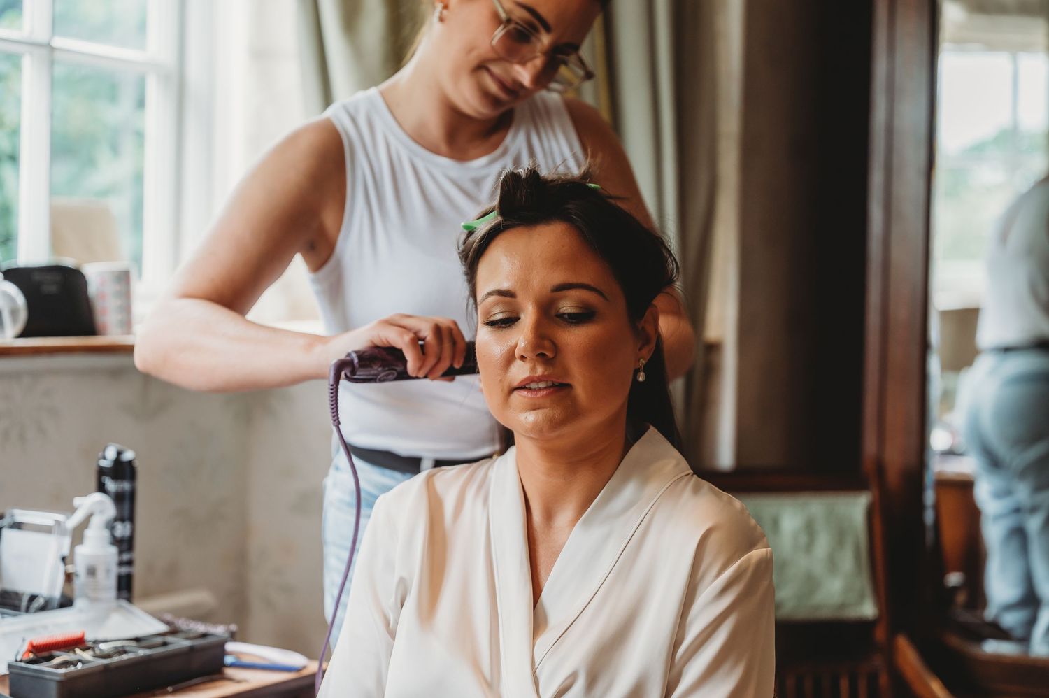 Stylist combs and styles hair of client wearing white robe during salon appointment in warm-lit setting.