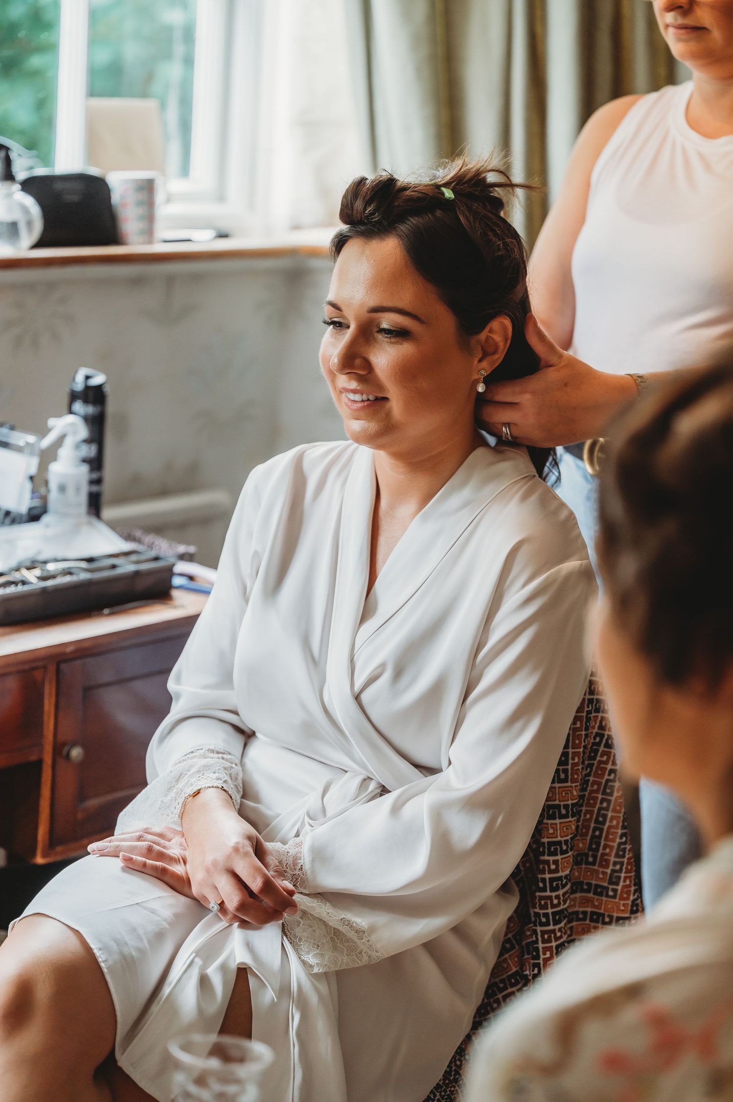 A smiling person in a white robe sits while having their hair styled during wedding preparation.