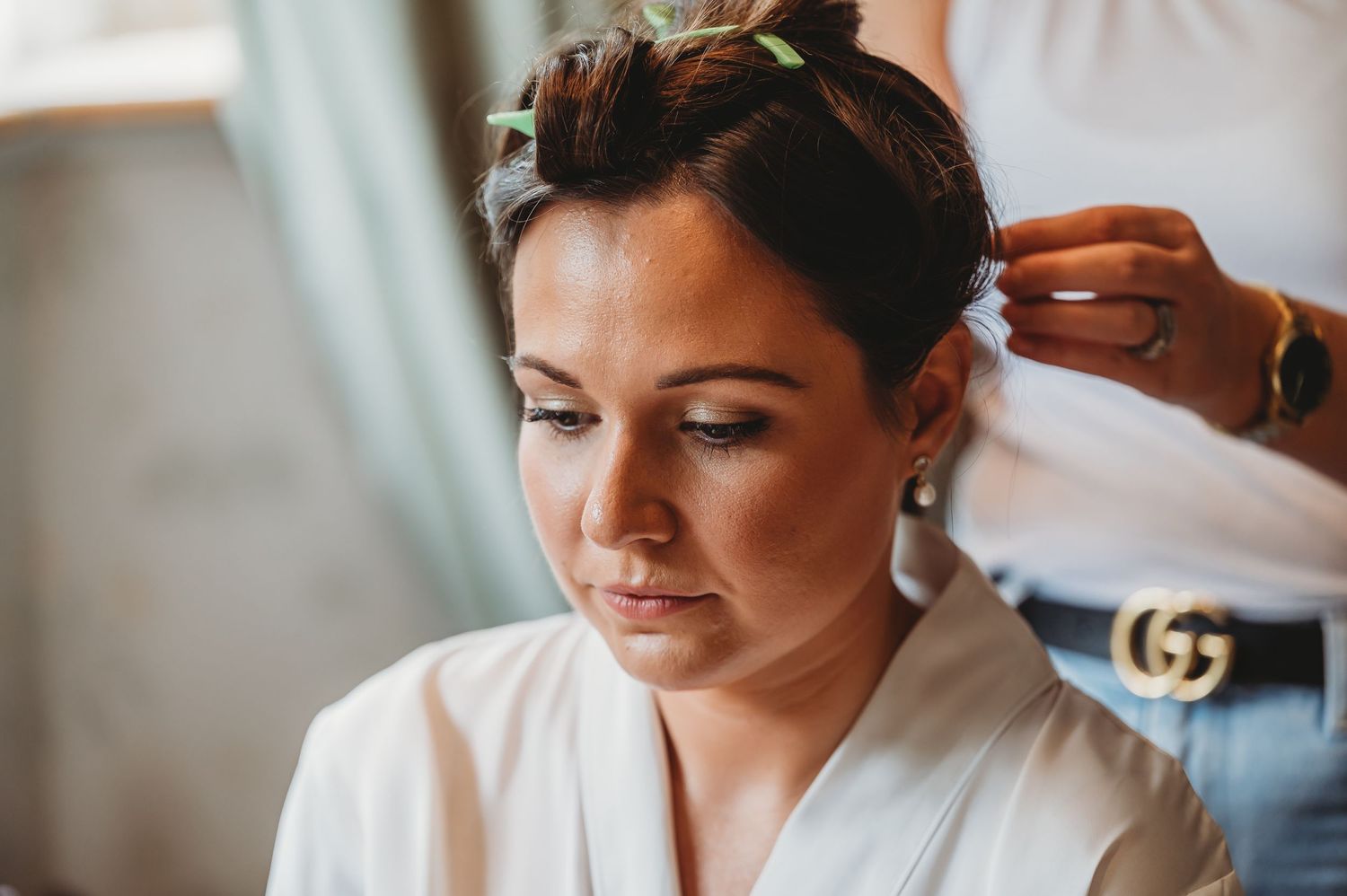 A hairstylist works on styling hair while the client in a white robe sits calmly with eyes closed.