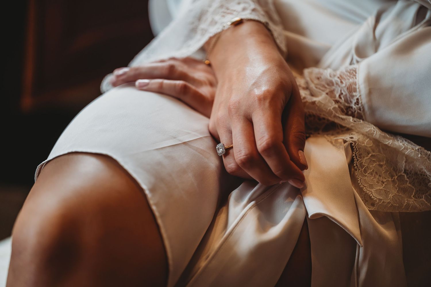Close-up shot of hands resting on white lace fabric and dress in warm dramatic lighting.