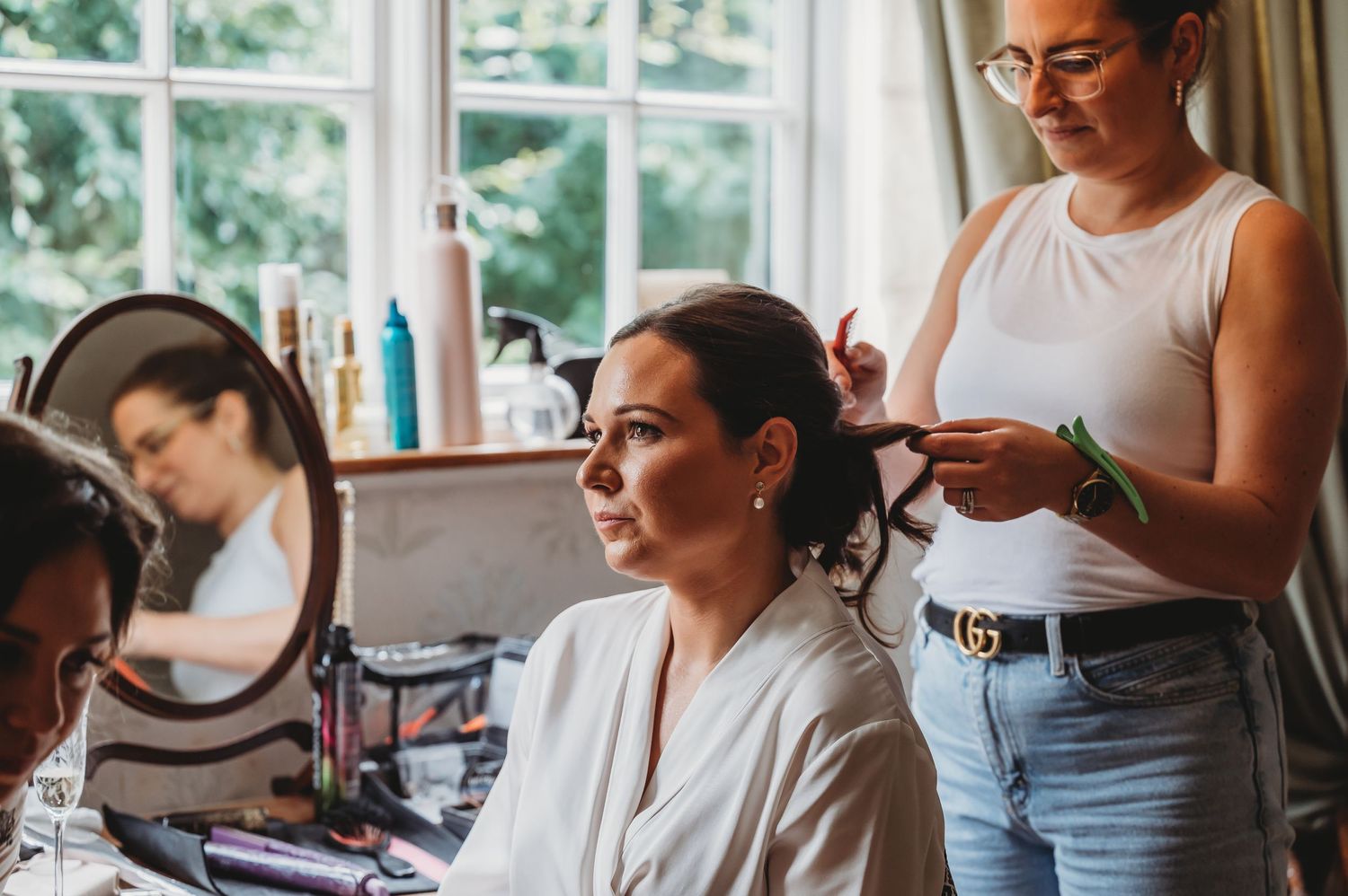 Hair stylist in blue jeans and white top styles bride's hair at vanity mirror during wedding preparation.
