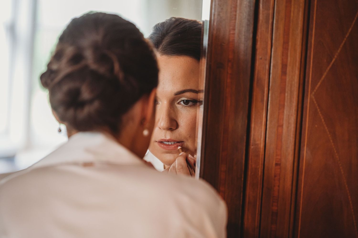 A woman looks at her reflection thoughtfully in a wooden framed mirror with soft natural lighting.