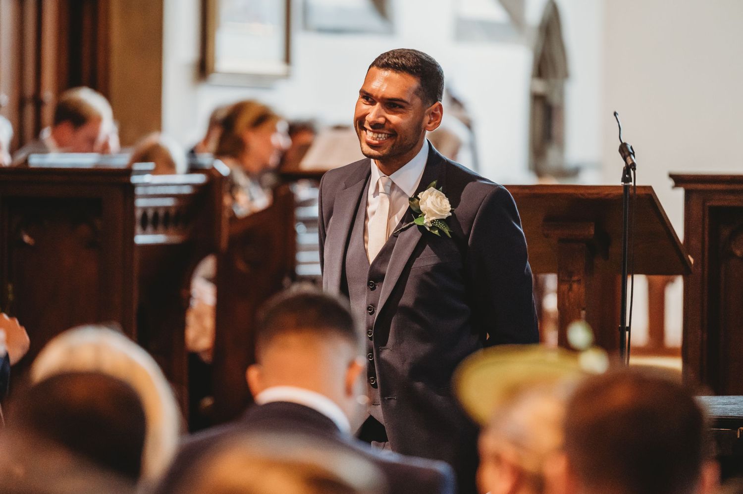 Groom smiling while standing at the altar in a grey suit during a church wedding ceremony.