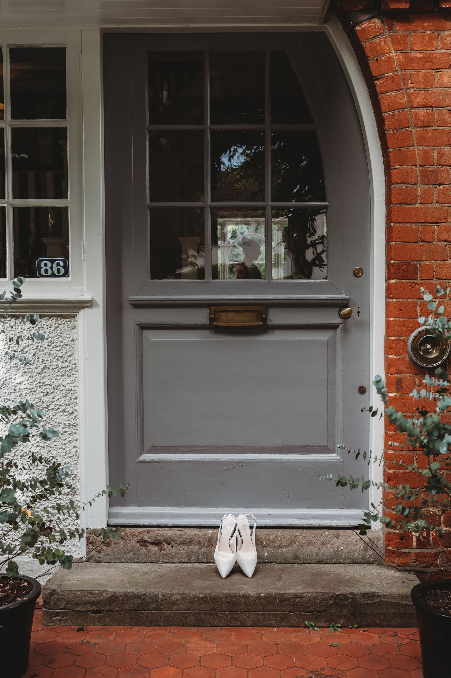 White wedding shoes sit on the doorstep of a grey arched door with glass panels and brick exterior details.