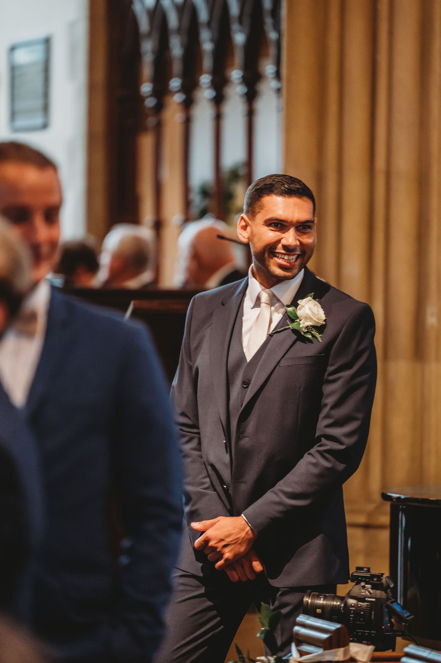 A joyful groom in a dark suit and tie smiles warmly at his wedding ceremony in an elegant church setting.