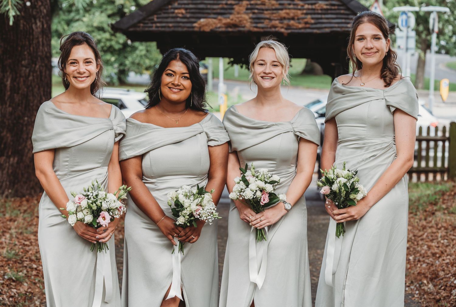 Four bridesmaids wearing matching light gray off-shoulder dresses stand together holding white floral bouquets outdoors.