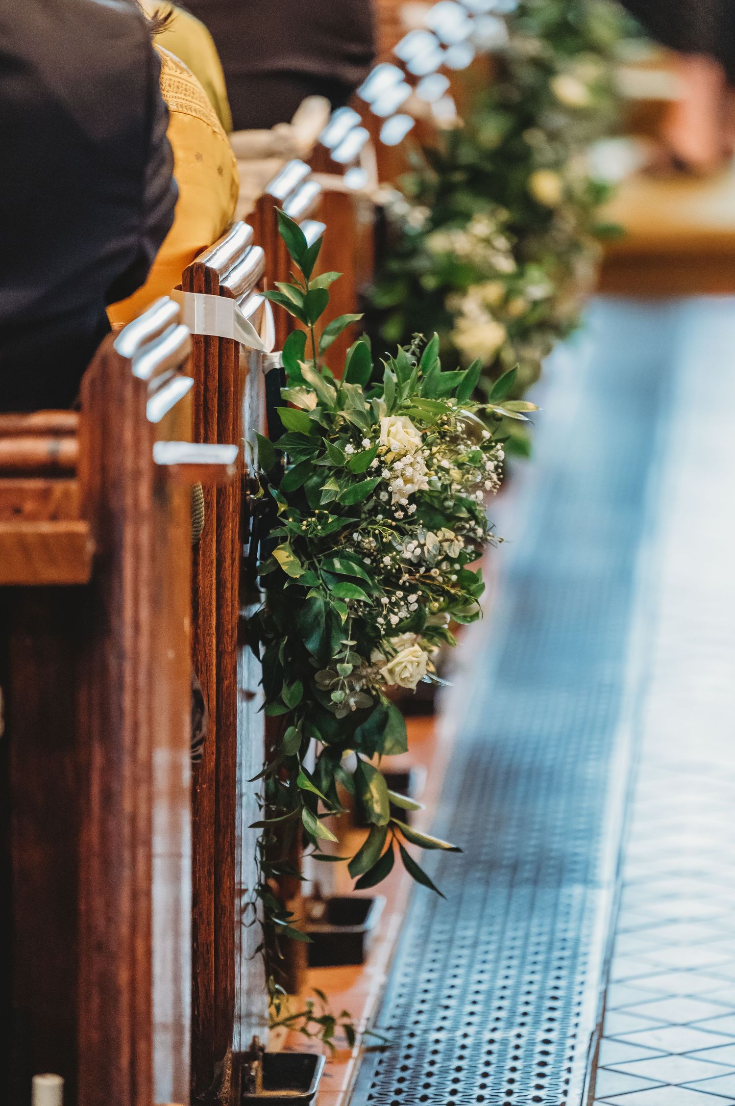 Elegant floral arrangements with green foliage decorate wooden church pews for a wedding ceremony.