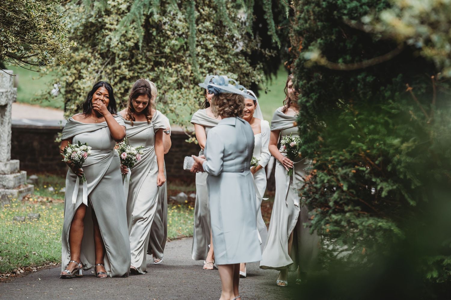 Wedding party in flowing silver and light blue dresses walks together on a garden path during outdoor celebration.