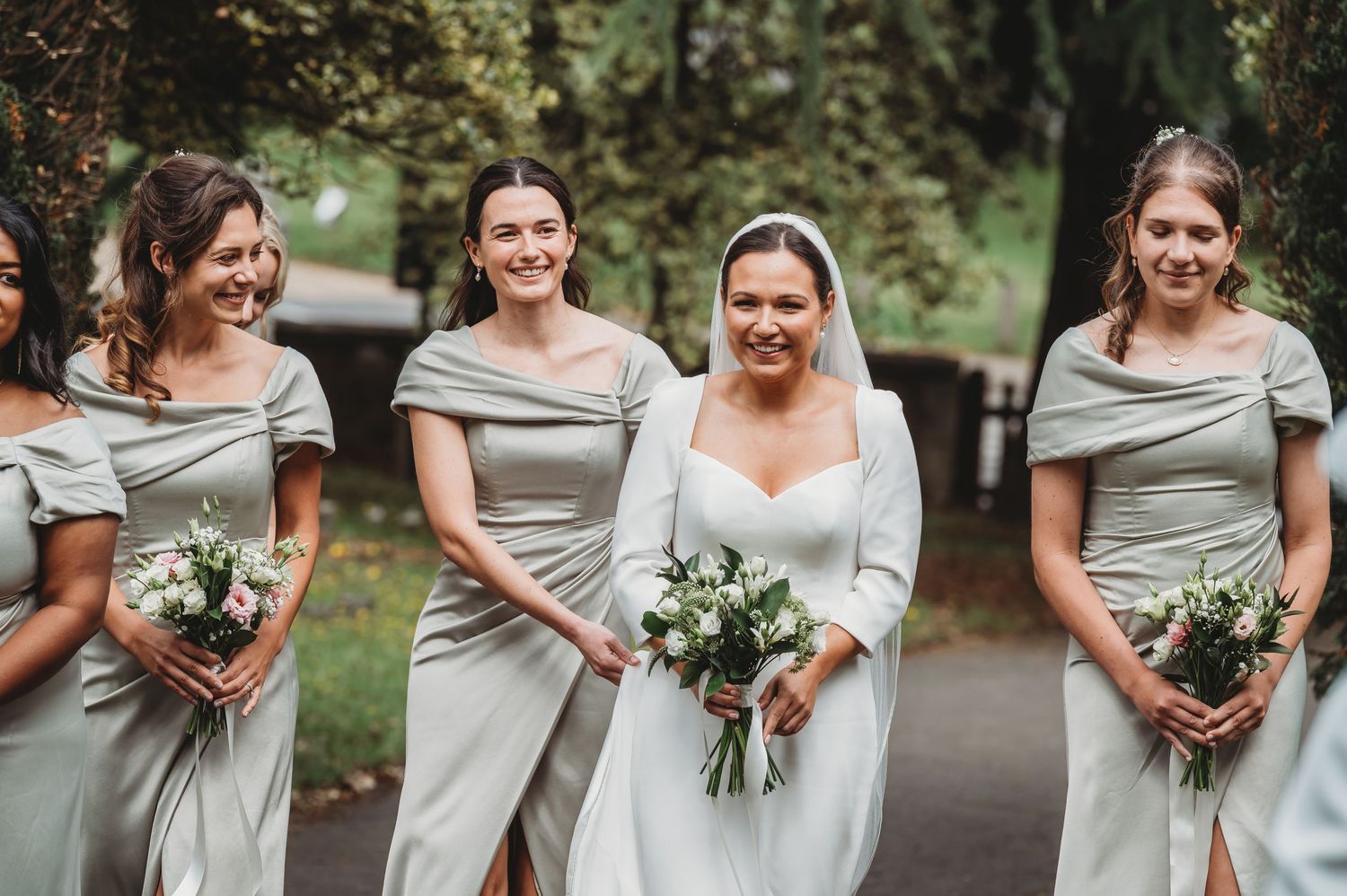 Wedding party in coordinated grey dresses holding floral bouquets on garden path.