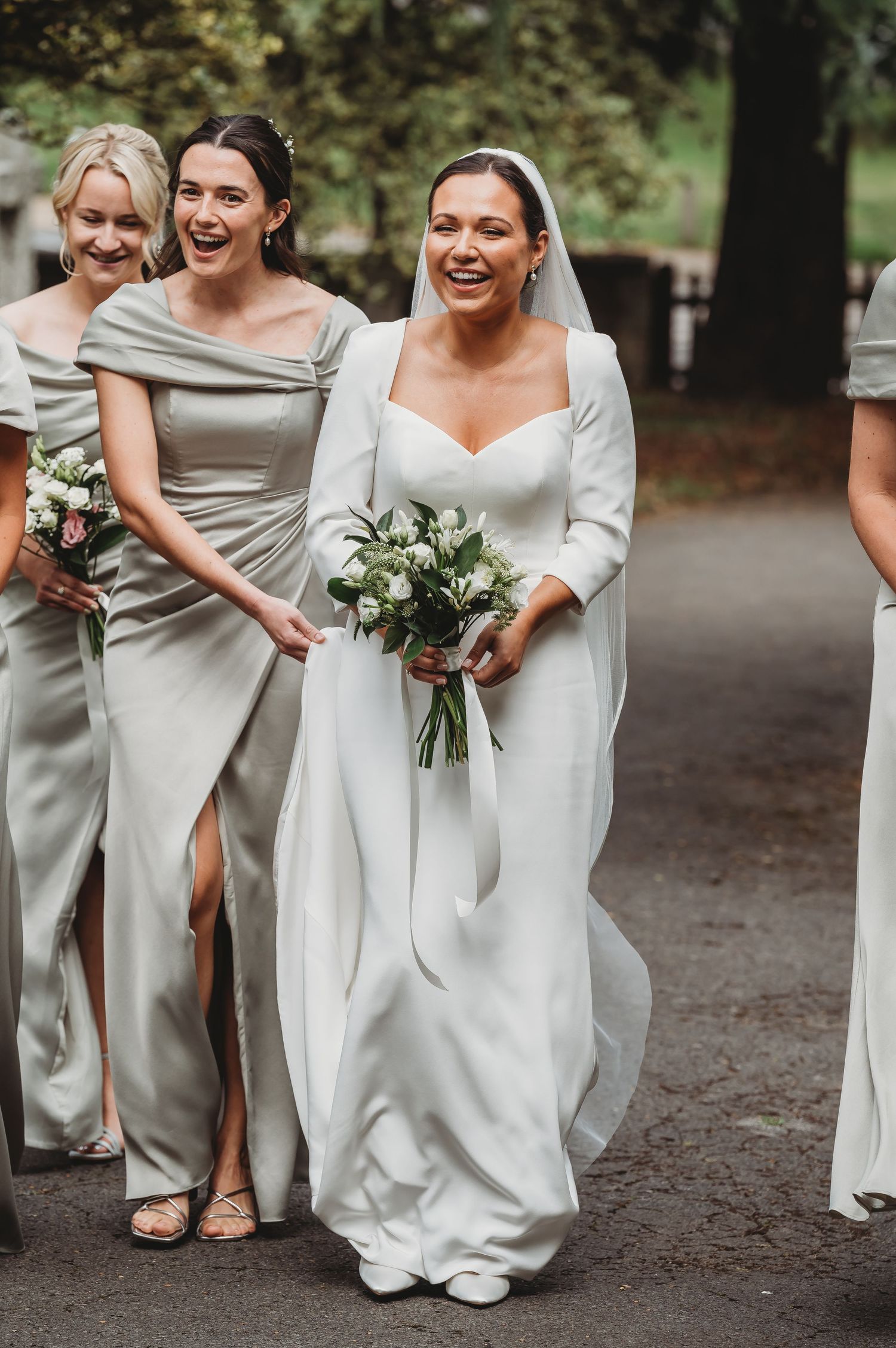 Bridal party in elegant grey dresses walking together outdoors holding bouquets.