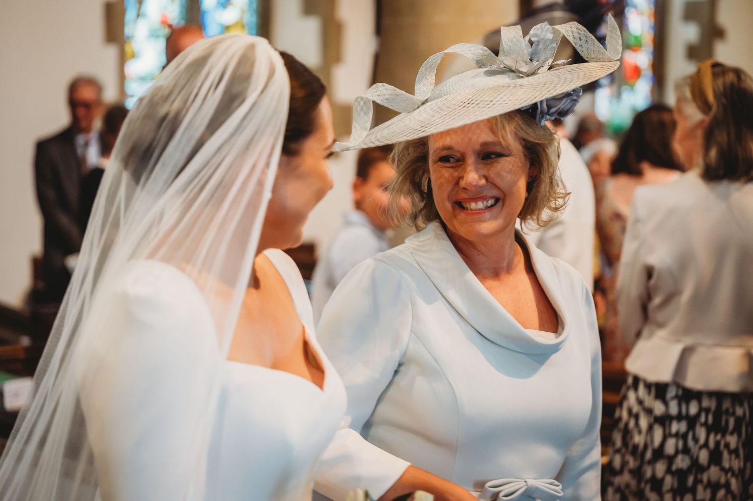 A joyful moment between a bride in a white wedding dress and veil and another wedding guest in a light blue outfit with hat.