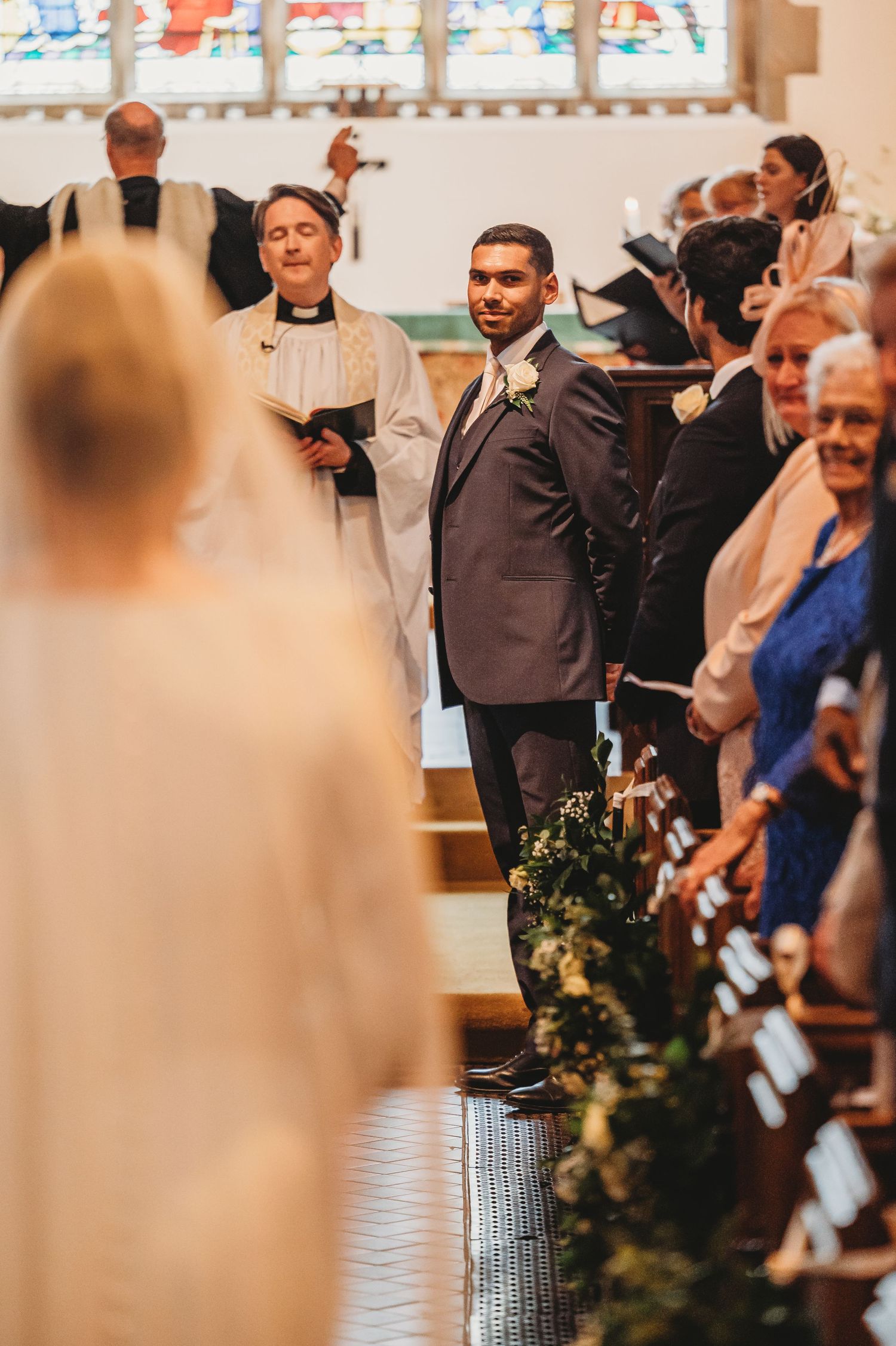 A wedding ceremony in a church with decorative greenery lining the aisle and guests seated in pews.