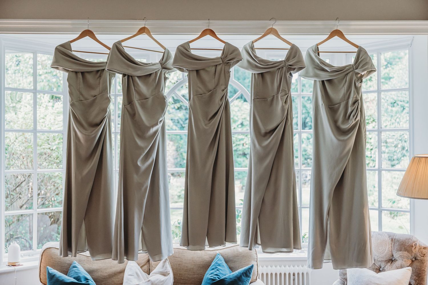 Five elegant taupe bridesmaid dresses hanging in a row on wooden hangers by a sunlit window with blue accent pillows below.