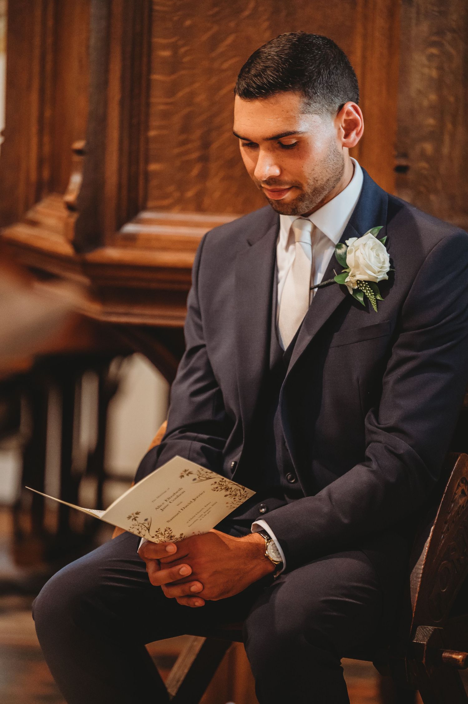 A groom in a navy suit with boutonniere reads from a program while seated in an elegant wooden church interior.