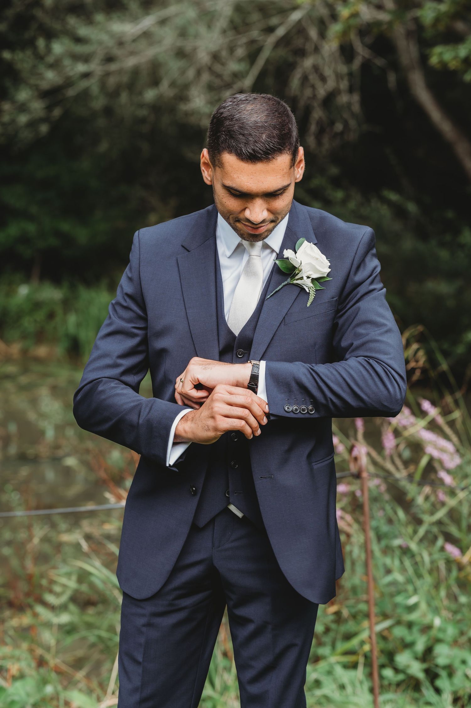 Groom in navy blue three-piece suit checking his watch while wearing a white boutonniere in outdoor garden setting.