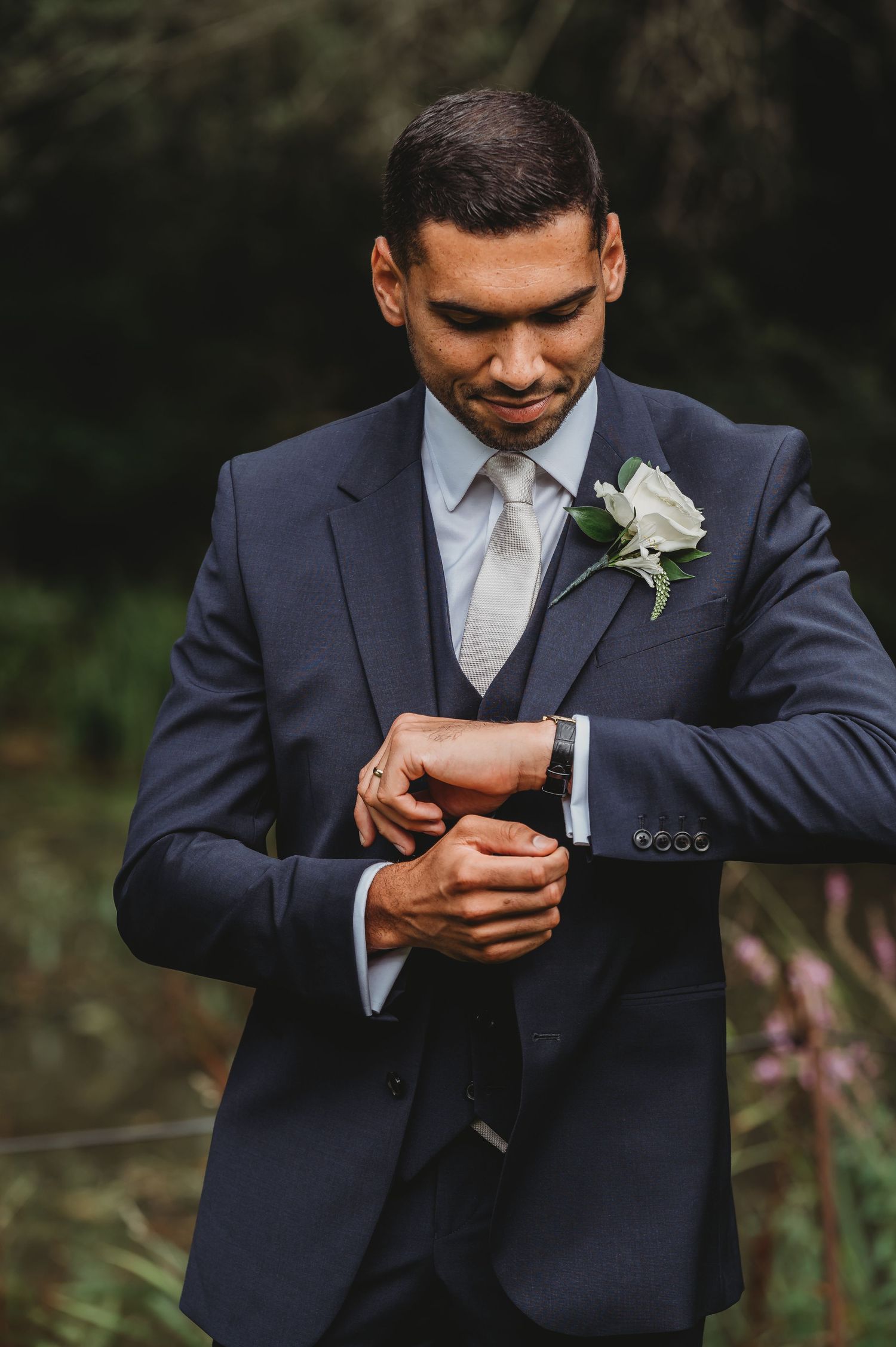 A groom in a navy blue suit and white boutonniere checks his watch during an outdoor wedding photoshoot.