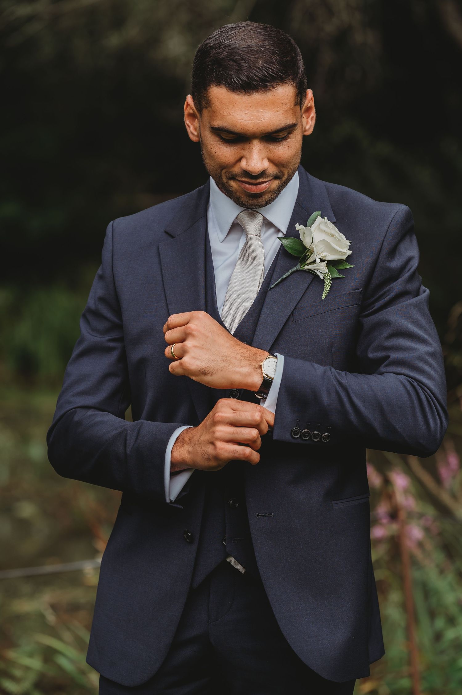 Groom in navy suit adjusts sleeves while wearing white boutonniere flower against dark outdoor background.