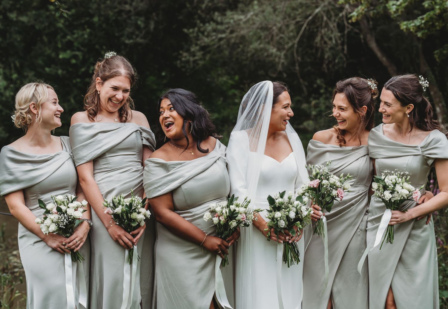 Bridesmaids in elegant sage grey dresses share a candid moment while holding white floral bouquets.