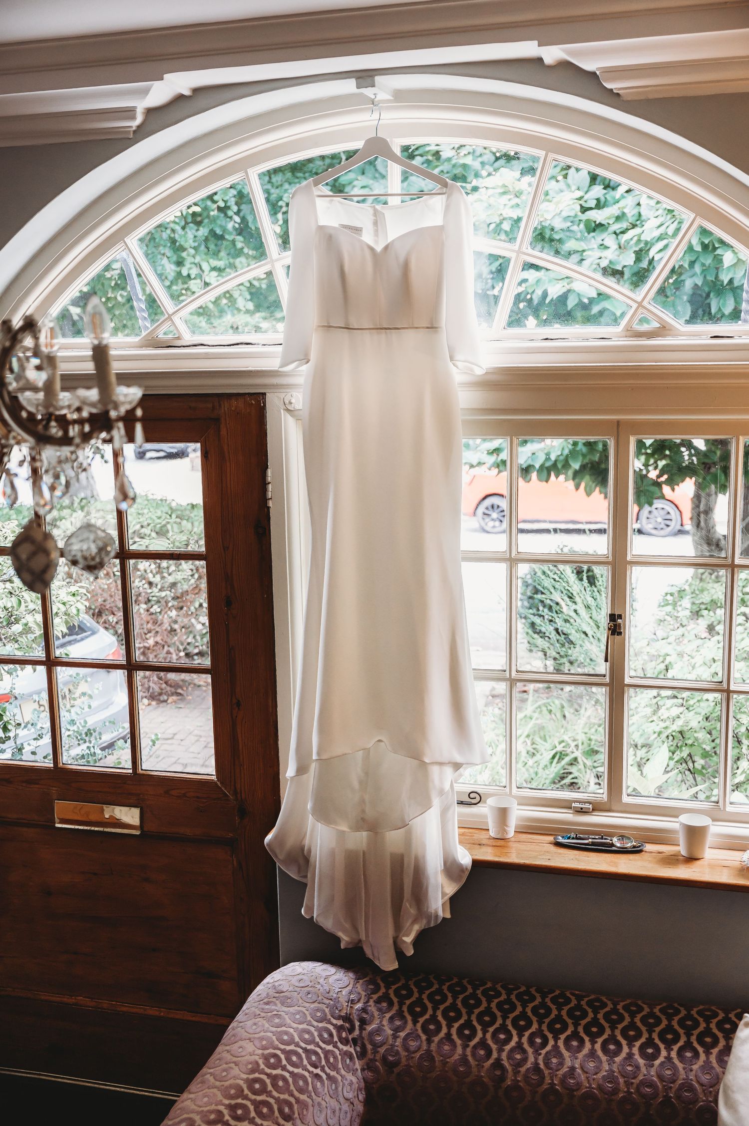 A white wedding dress hangs elegantly in front of an arched window with natural light streaming through in a rustic interior.