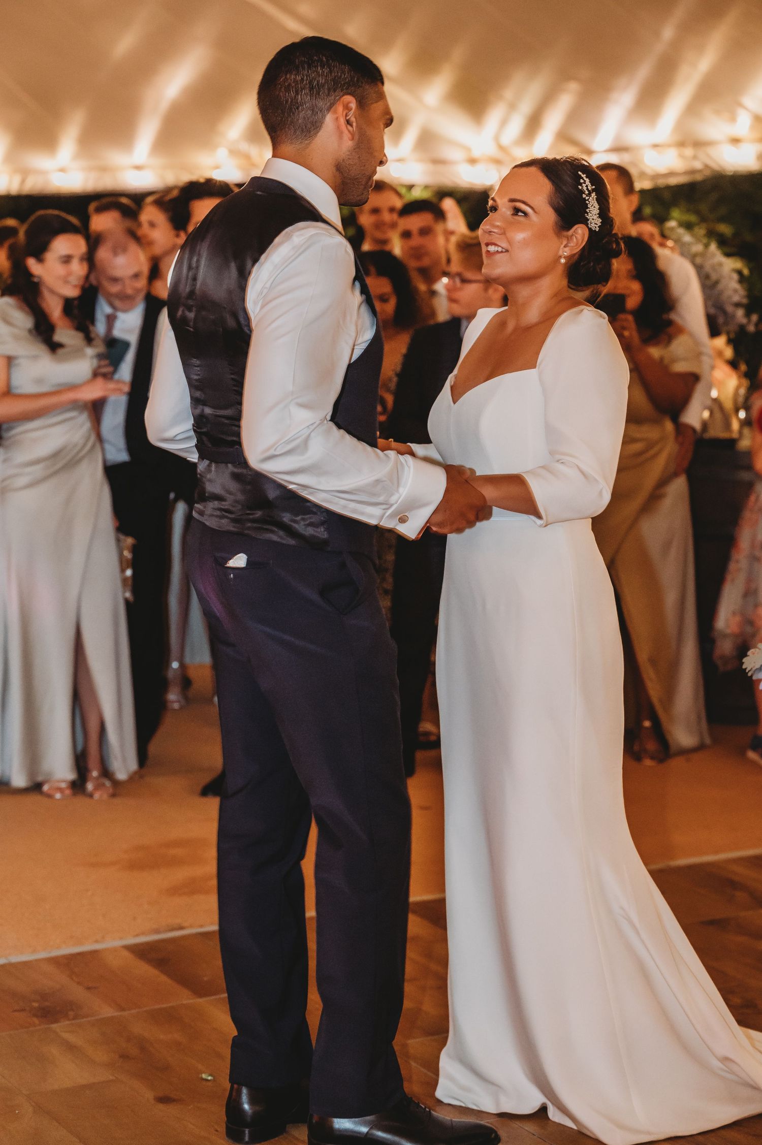 A couple dances their first dance at their wedding reception under a tent with guests watching in the background.