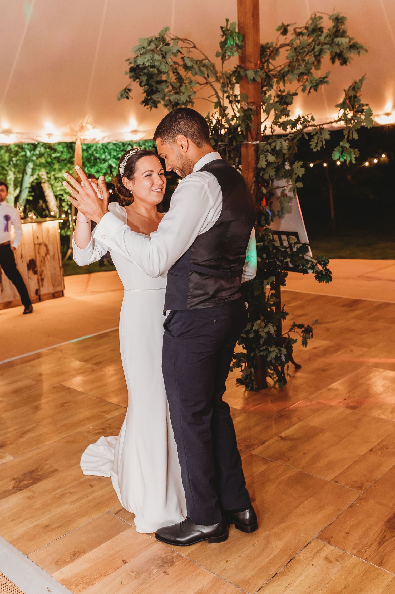 A couple shares their first dance as newlyweds on a wooden dance floor under twinkling lights and greenery decor.
