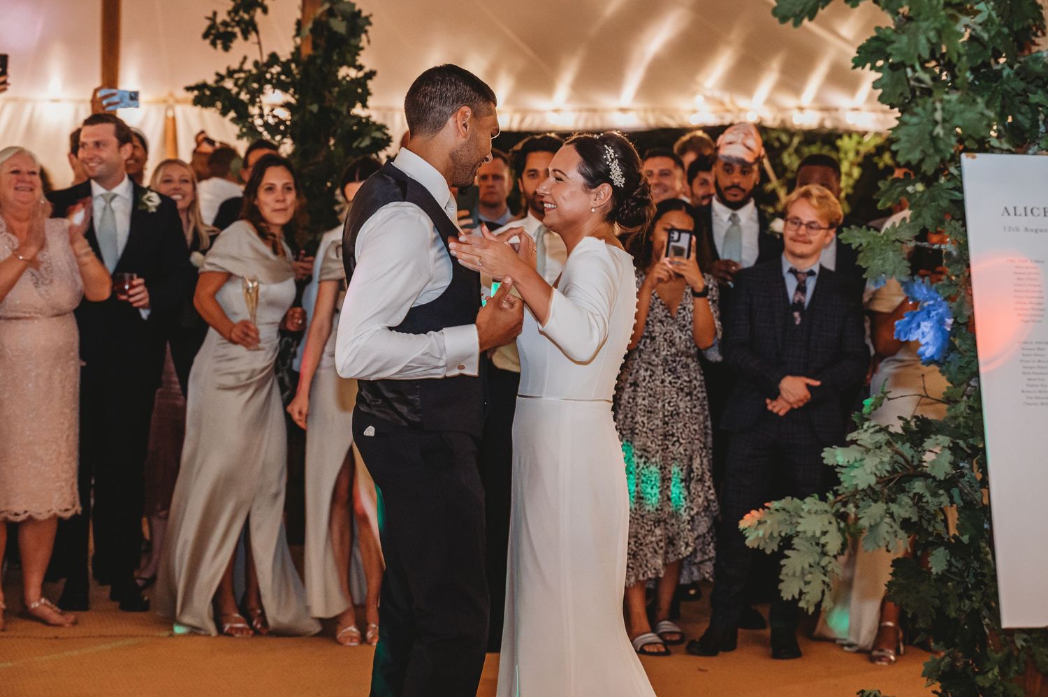 Couple shares their first dance surrounded by wedding guests under warm lighting in a decorated reception venue.