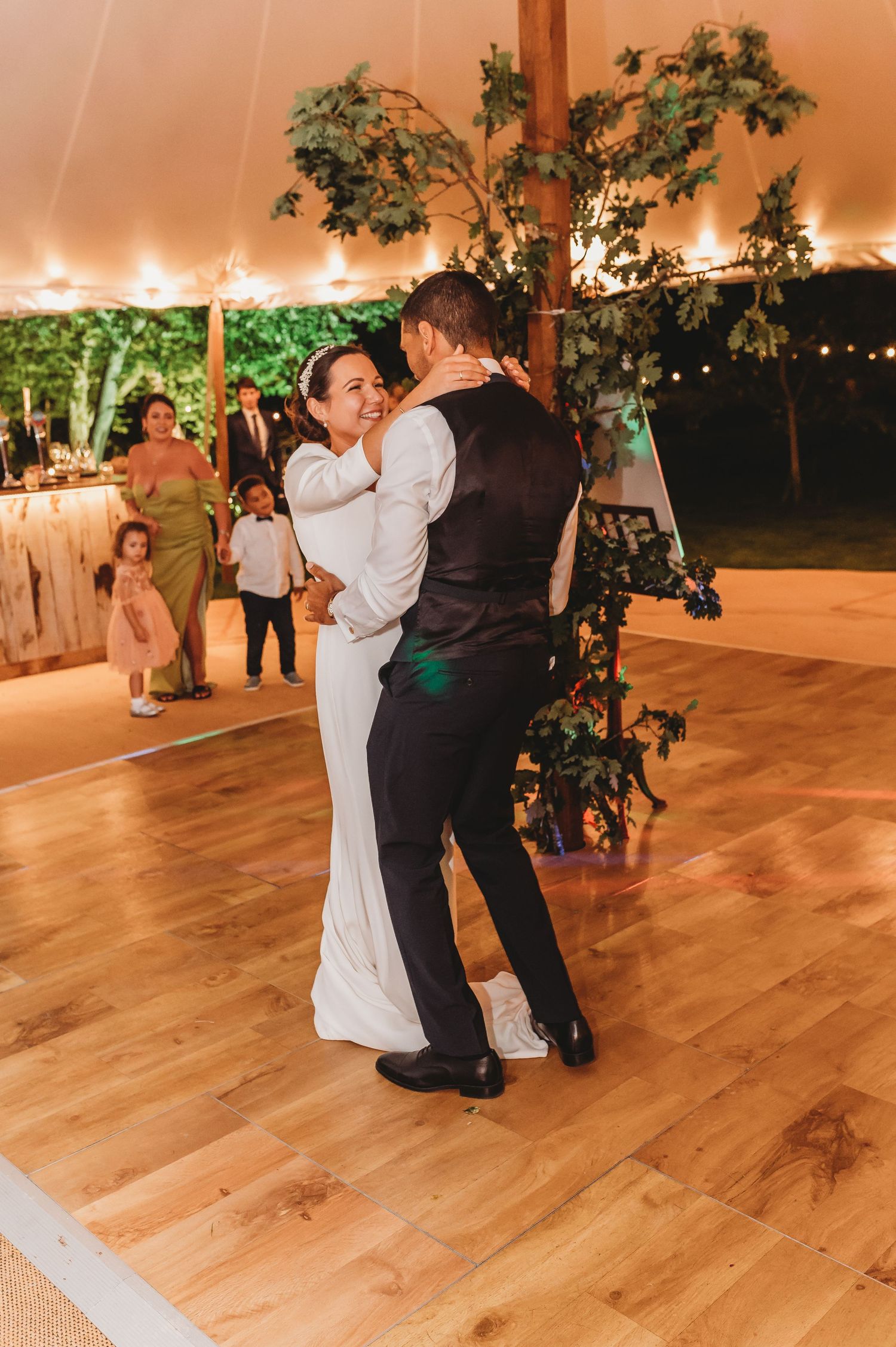 A couple shares an intimate dance at their wedding reception under a tent with twinkling lights and greenery decor.