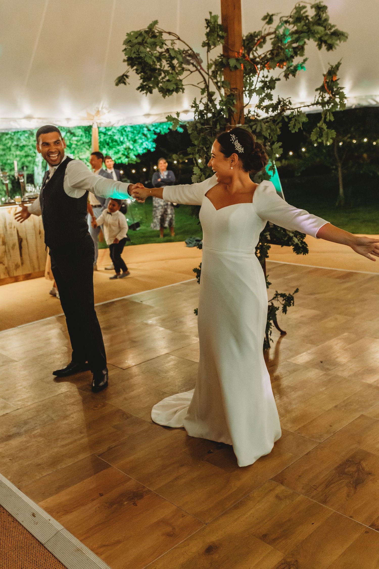 A couple dances together at their wedding reception on a wooden dance floor under a white tent with green lighting.