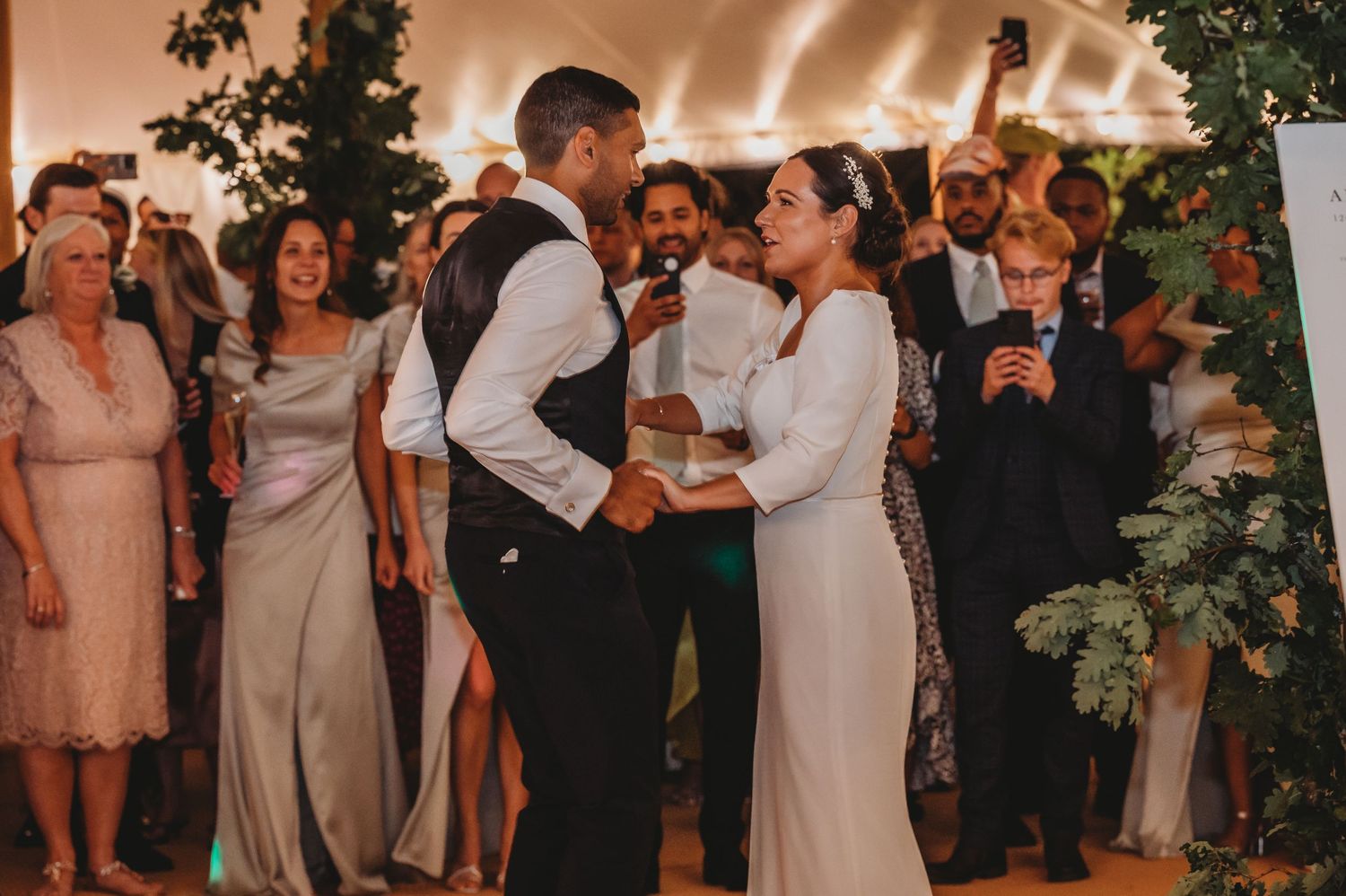 A couple sharing their first dance at a wedding reception while guests watch under warm string lights and floral decor.
