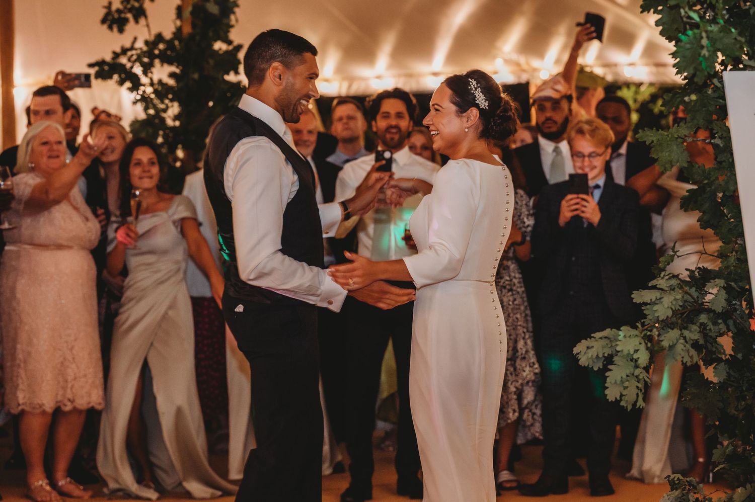 A couple exchanges rings during an intimate wedding ceremony under string lights while guests watch from the background.