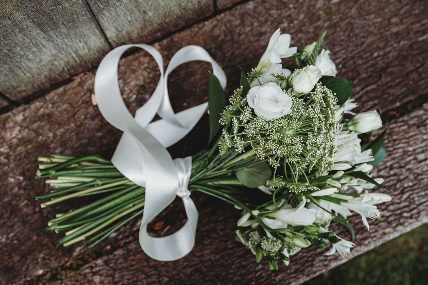A white wedding bouquet with fresh greenery and roses tied with an elegant white satin ribbon rests on wooden surface.