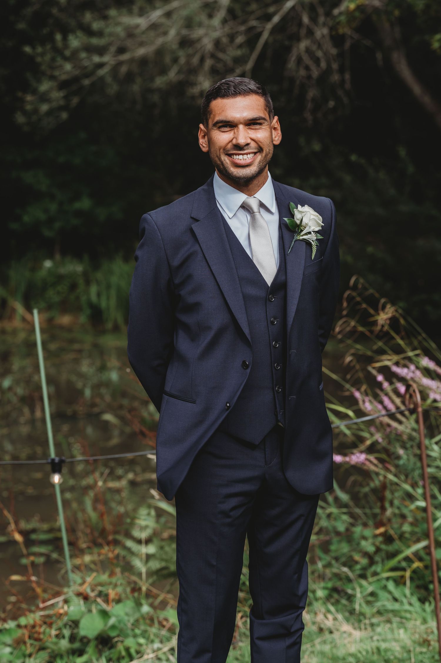 A smiling groom in a navy blue three-piece suit with gray tie and white boutonniere poses outdoors against greenery.