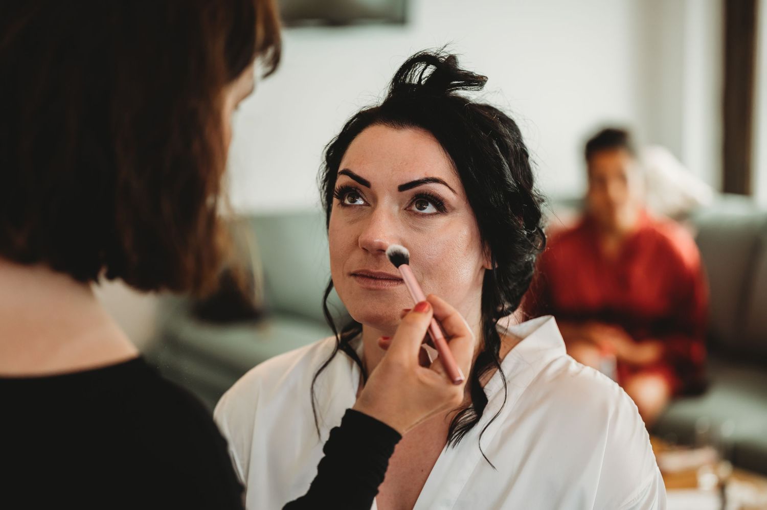 Makeup artist applies lip product on client while man sits blurred in background.
