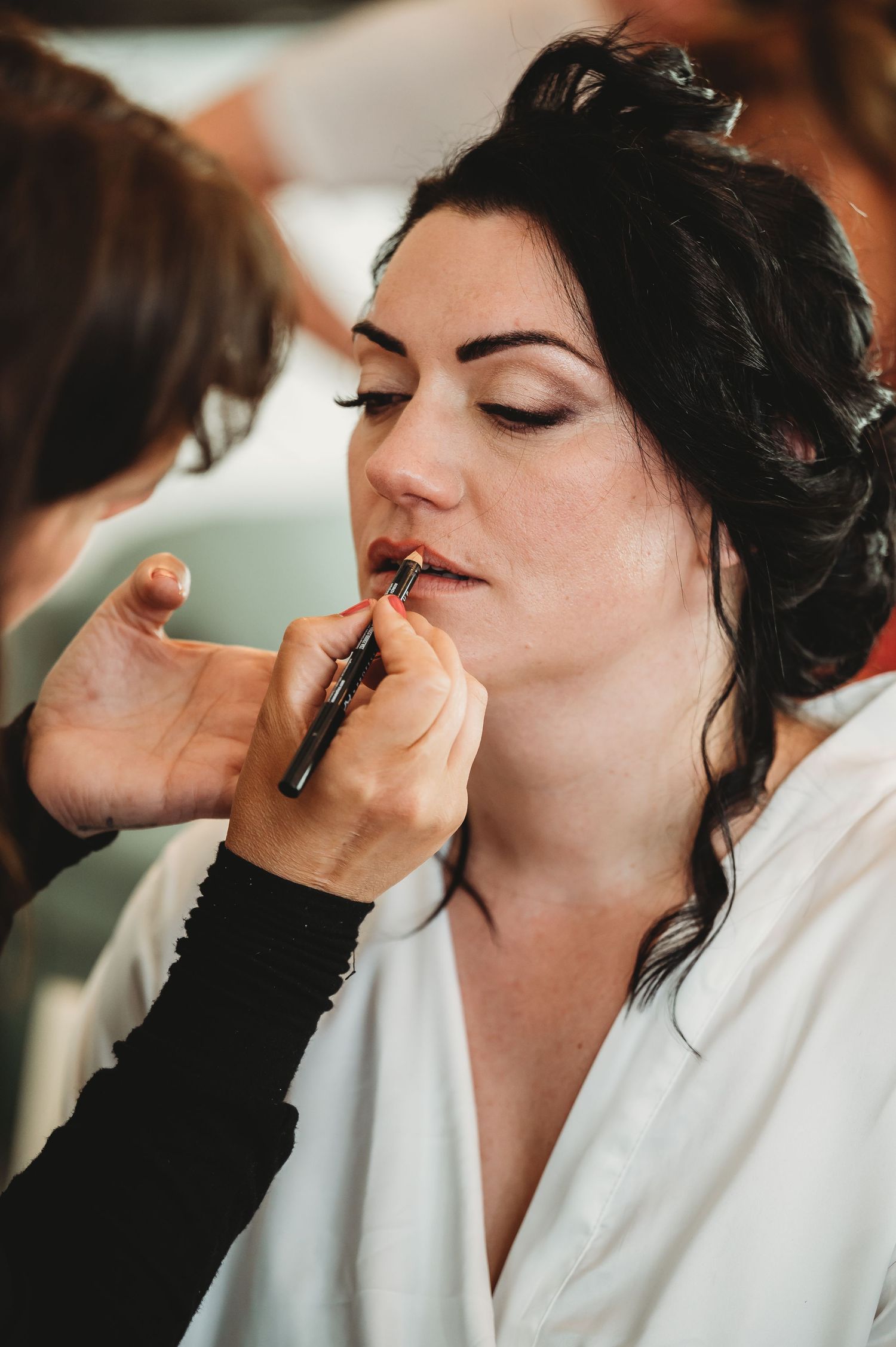 A makeup artist applies lipstick during a bridal preparation session with soft natural lighting.