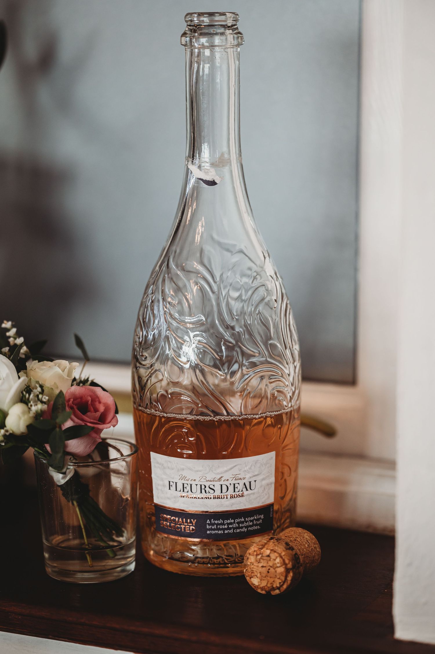 A bottle of rose wine with ornate glass pattern sits beside flowers and cork on wooden surface in moody lighting.