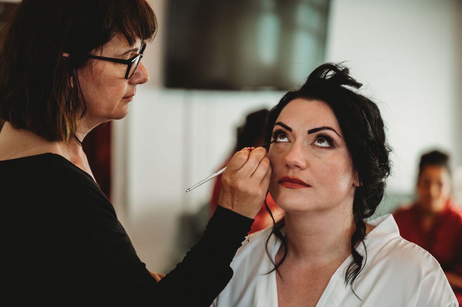 Professional makeup artist applies eye makeup to someone wearing a white robe during preparation for an event.