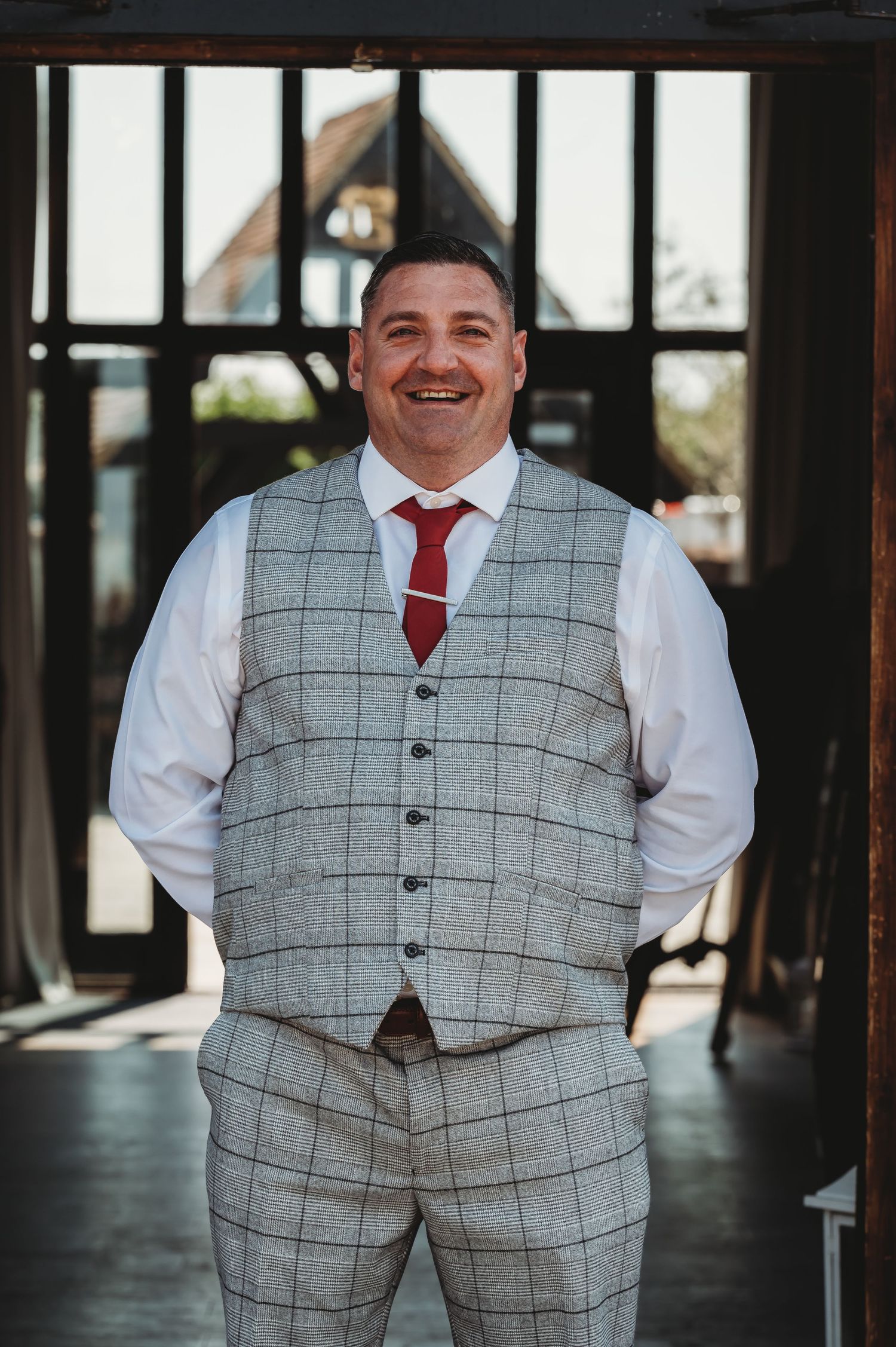 A person wearing a gray plaid vest and red tie stands smiling in front of a window on their wedding day.