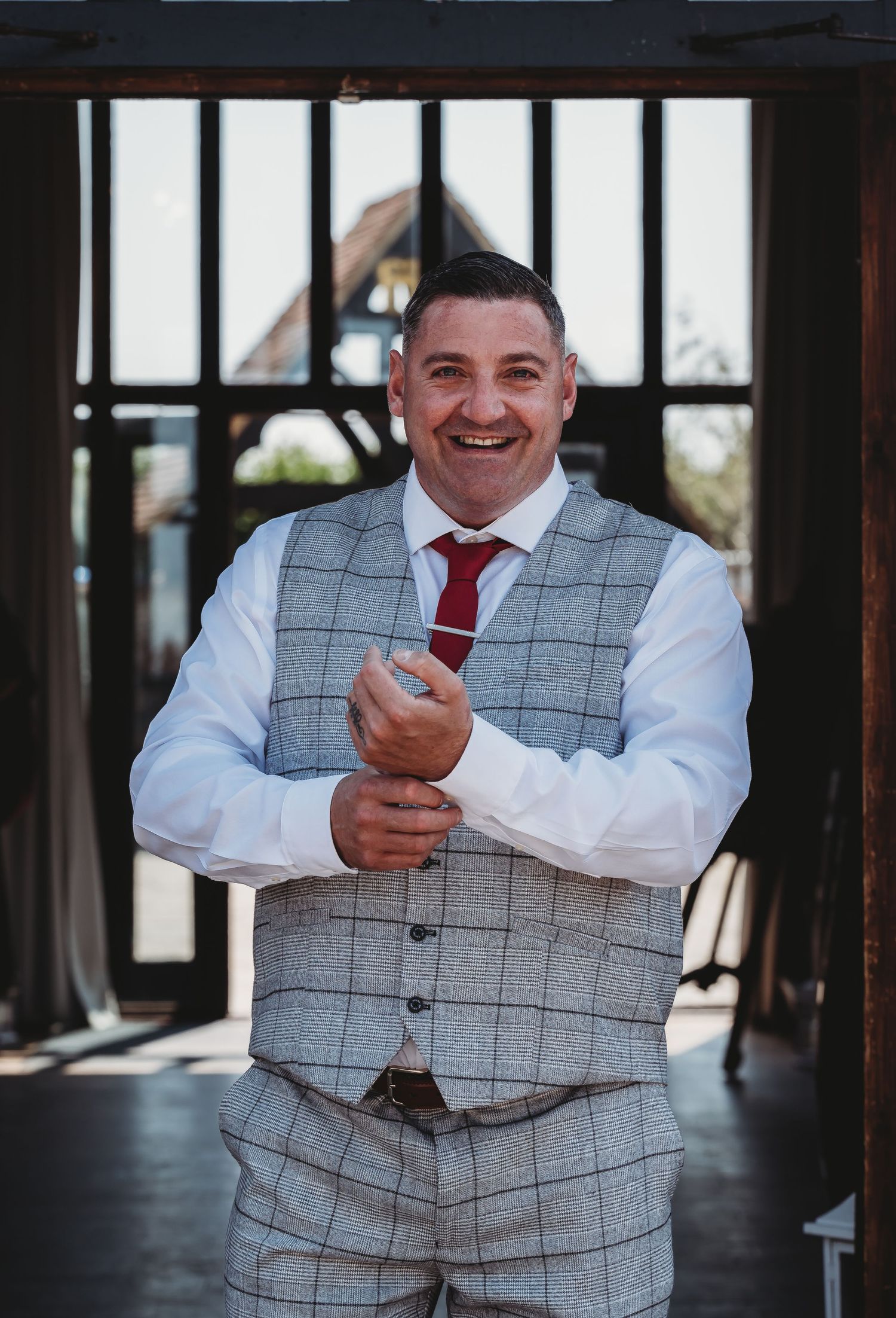 A person wearing a grey plaid vest and red tie stands with crossed arms in a rustic doorway.
