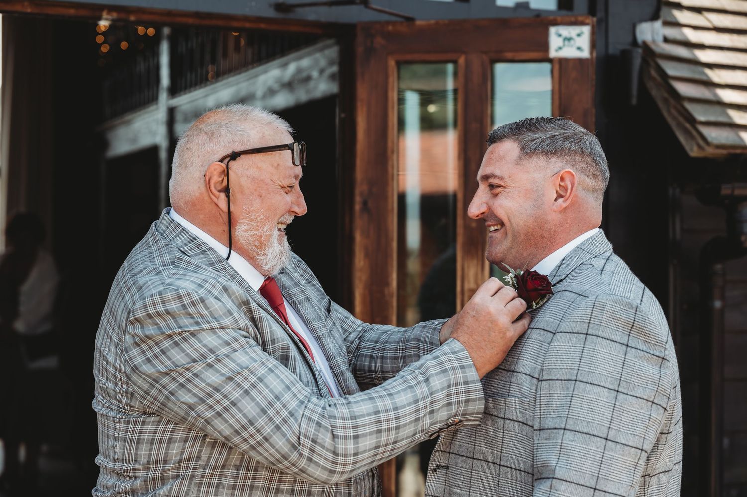 Two men in matching plaid blazers stand outside while one adjusts the other's tie with a smile.
