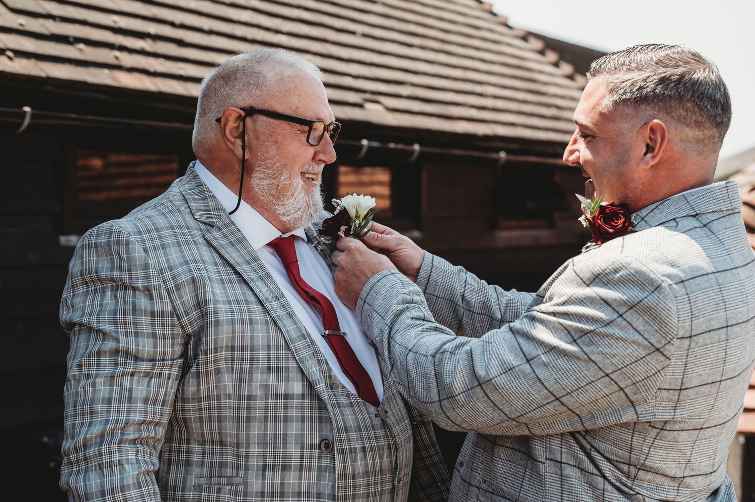 A groomsman in a light grey suit helps adjust the boutonniere on the groom's windowpane plaid suit.