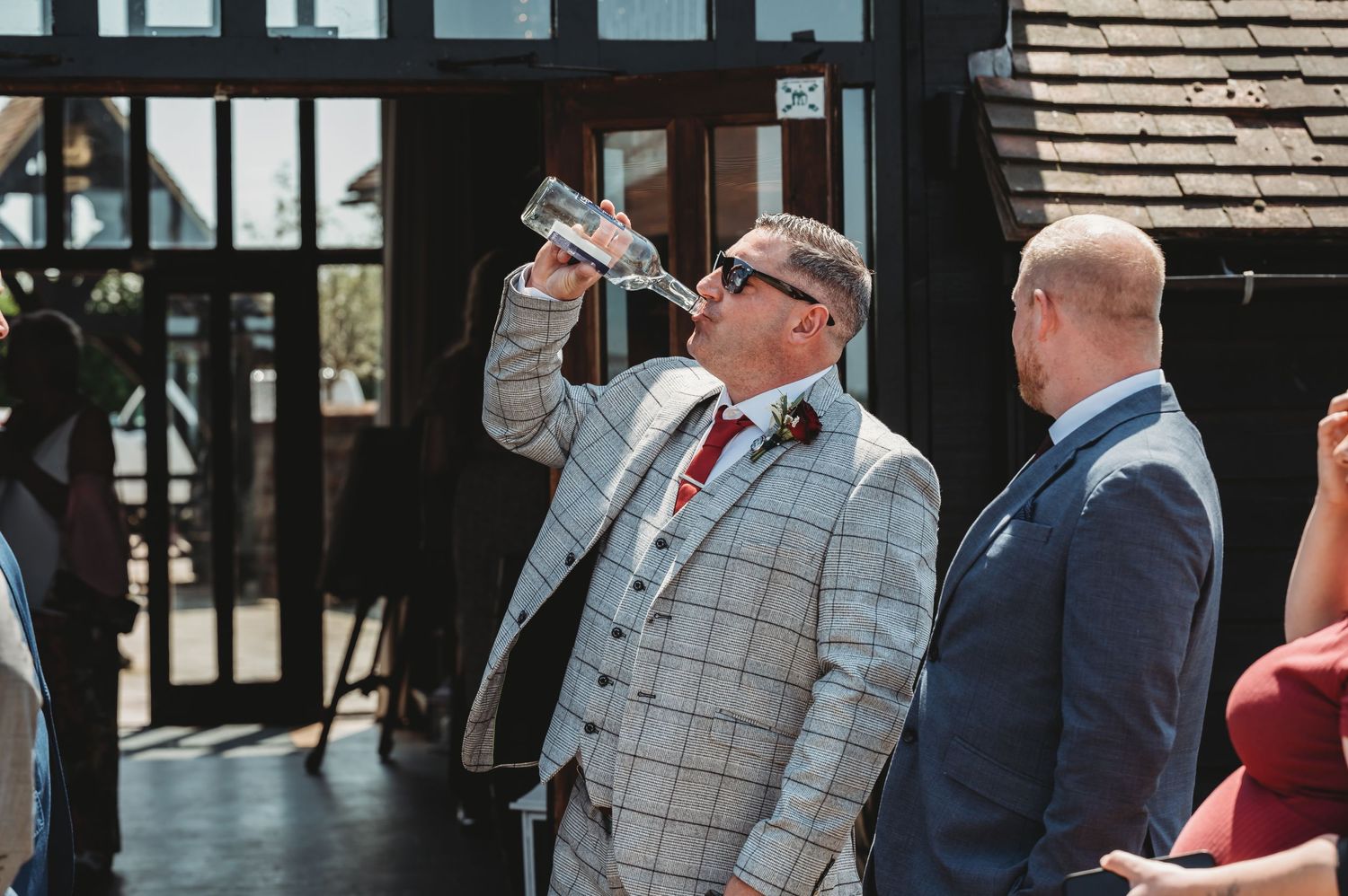 A guest in a gray checkered suit takes a drink during an outdoor celebration.