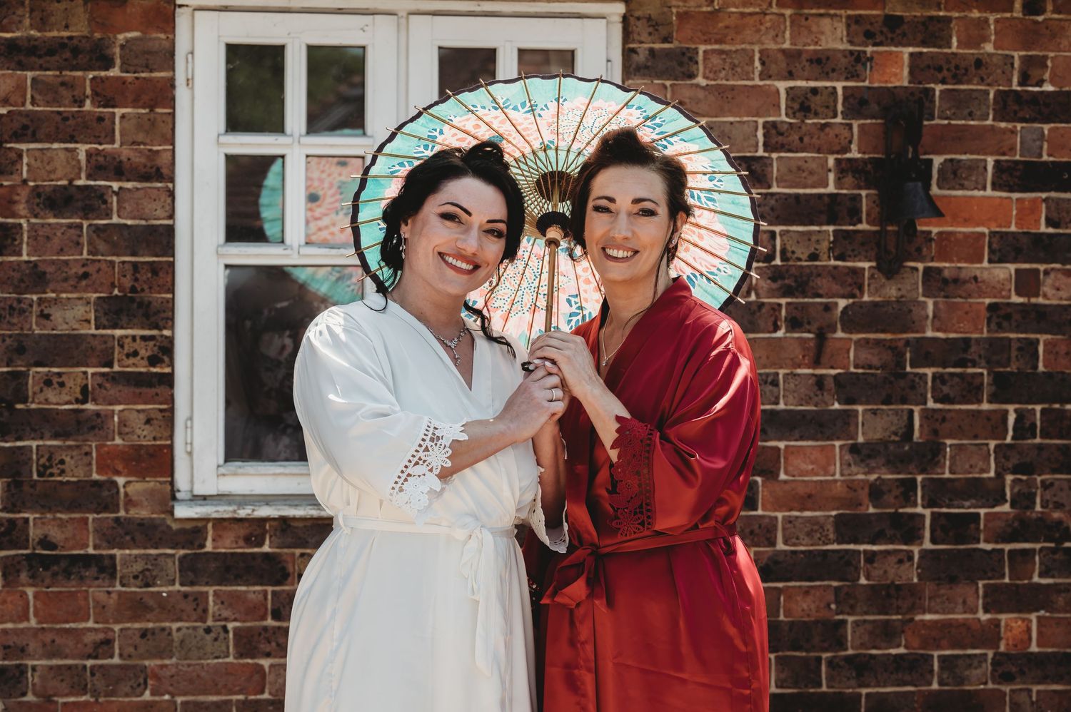 Two people in robes share an Asian paper parasol while posing against a rustic brick wall with a window.