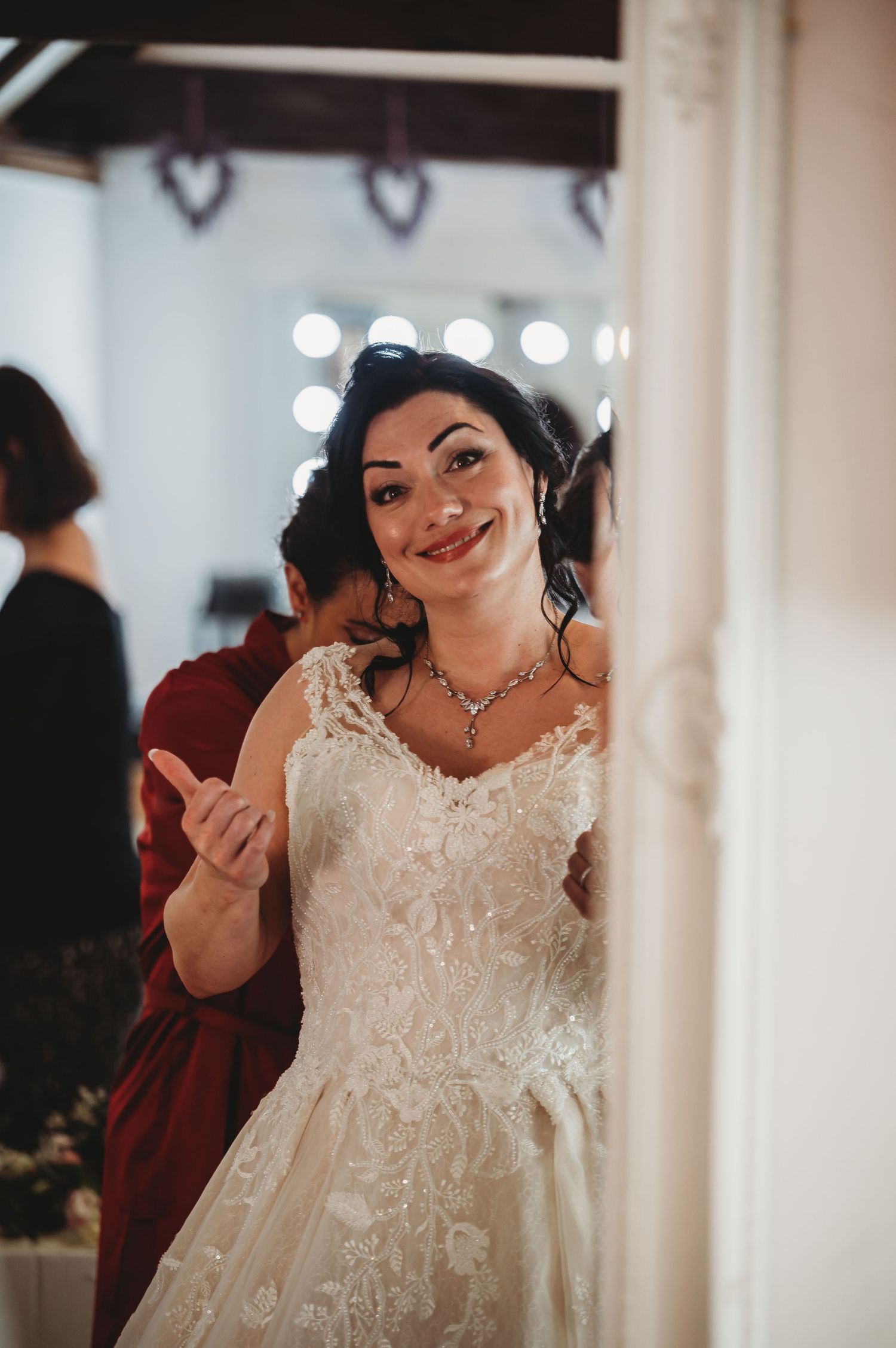 A bride wearing a white lace wedding dress and necklace smiles warmly in front of vanity lights.