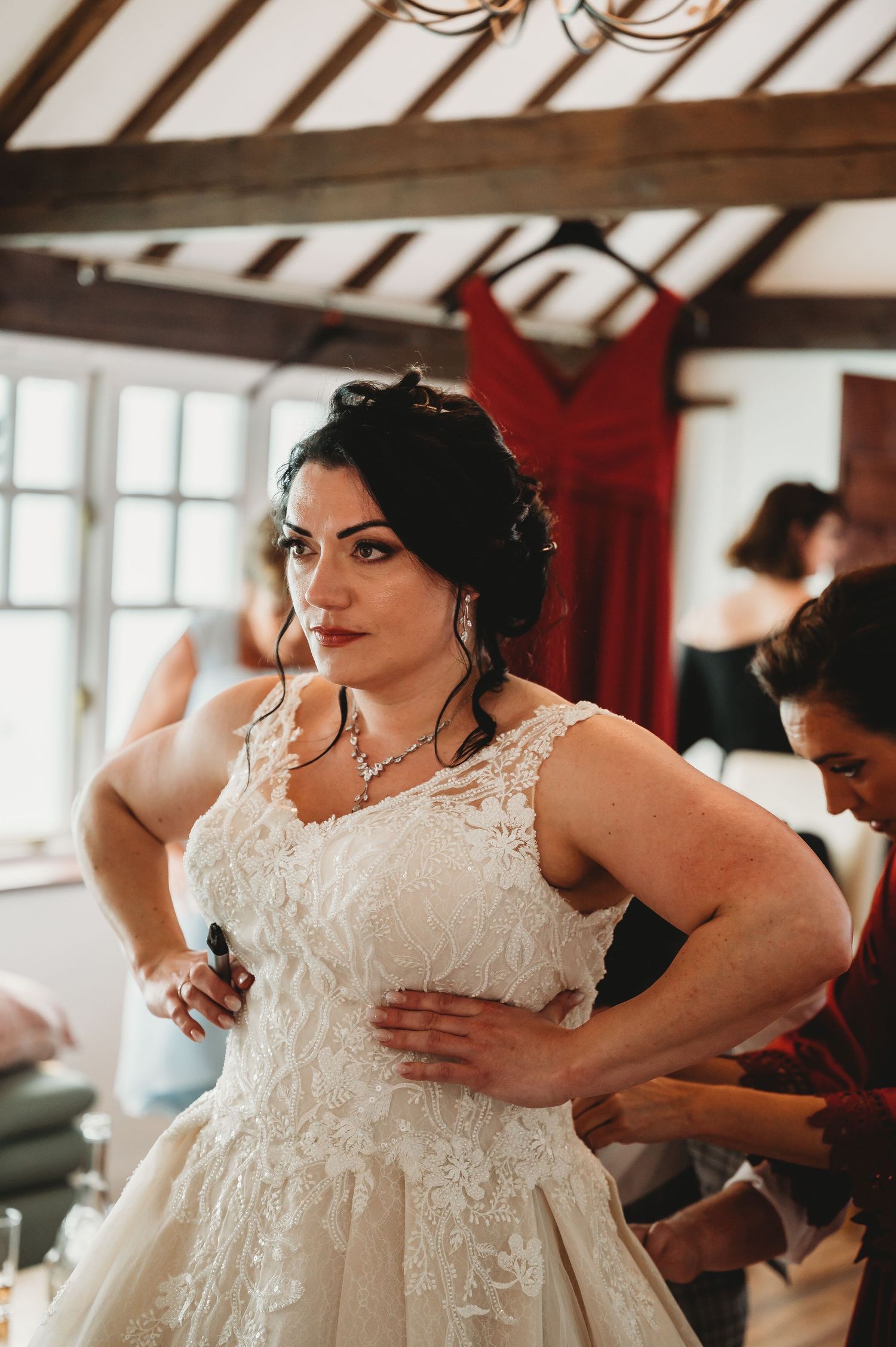 A bride in a white lace wedding dress gets ready in a room with exposed wooden beams and a red dress hanging behind her.
