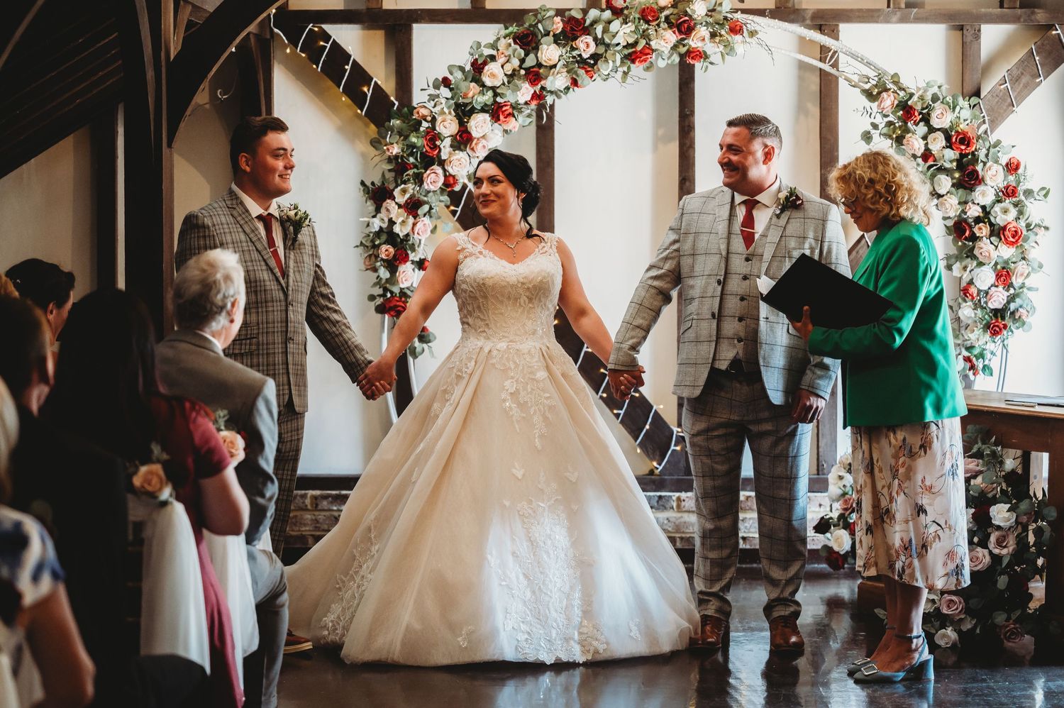 Wedding ceremony under a floral arch with people gathered around in gray and white attire.