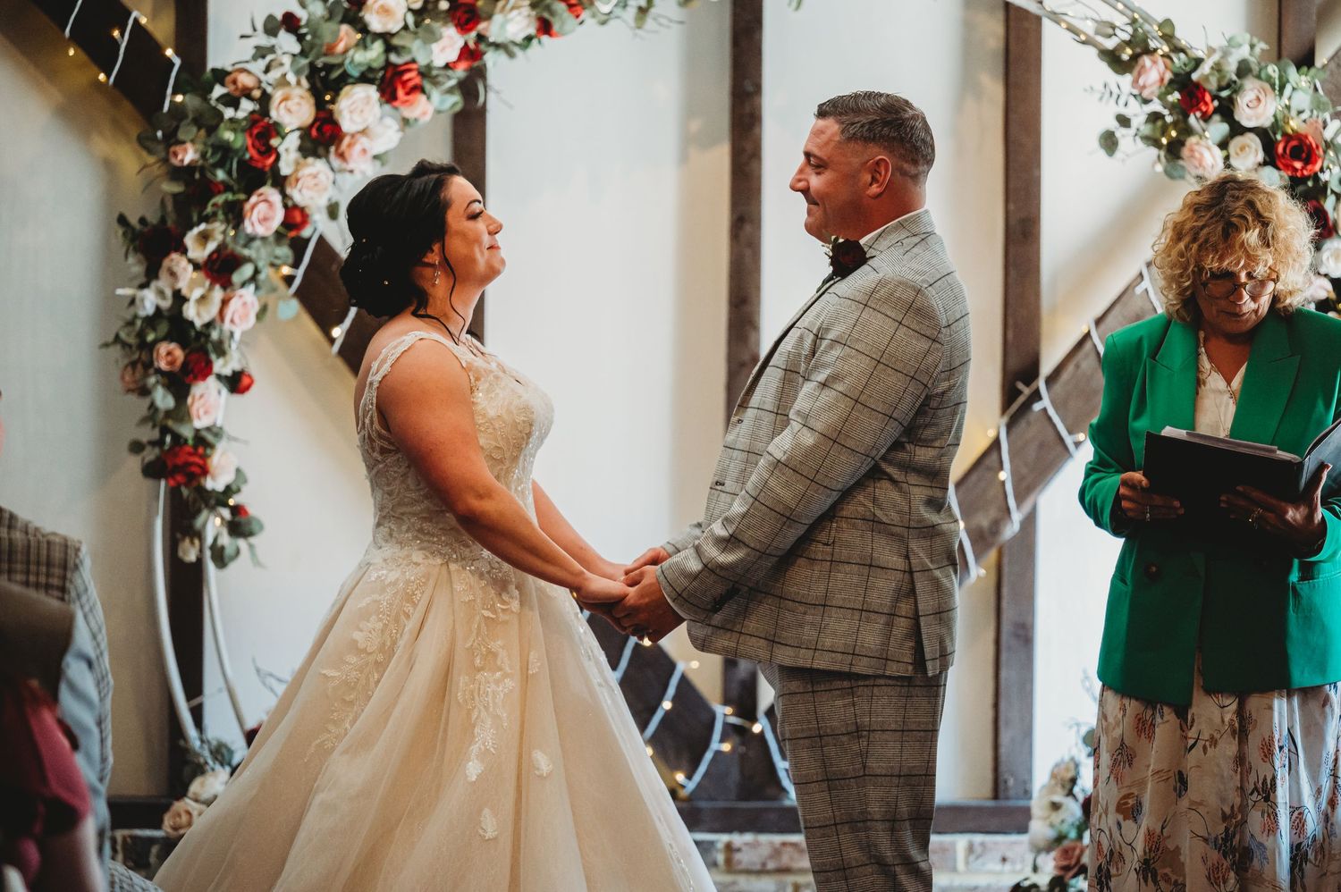 A couple holds hands at their wedding ceremony under a floral arch as an officiant in green reads from a book.